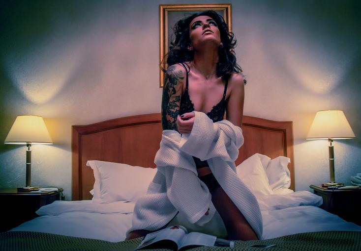 Žena s čiernymi vlasmi a tetovaním na ramene kľačí na posteli zahalená županom